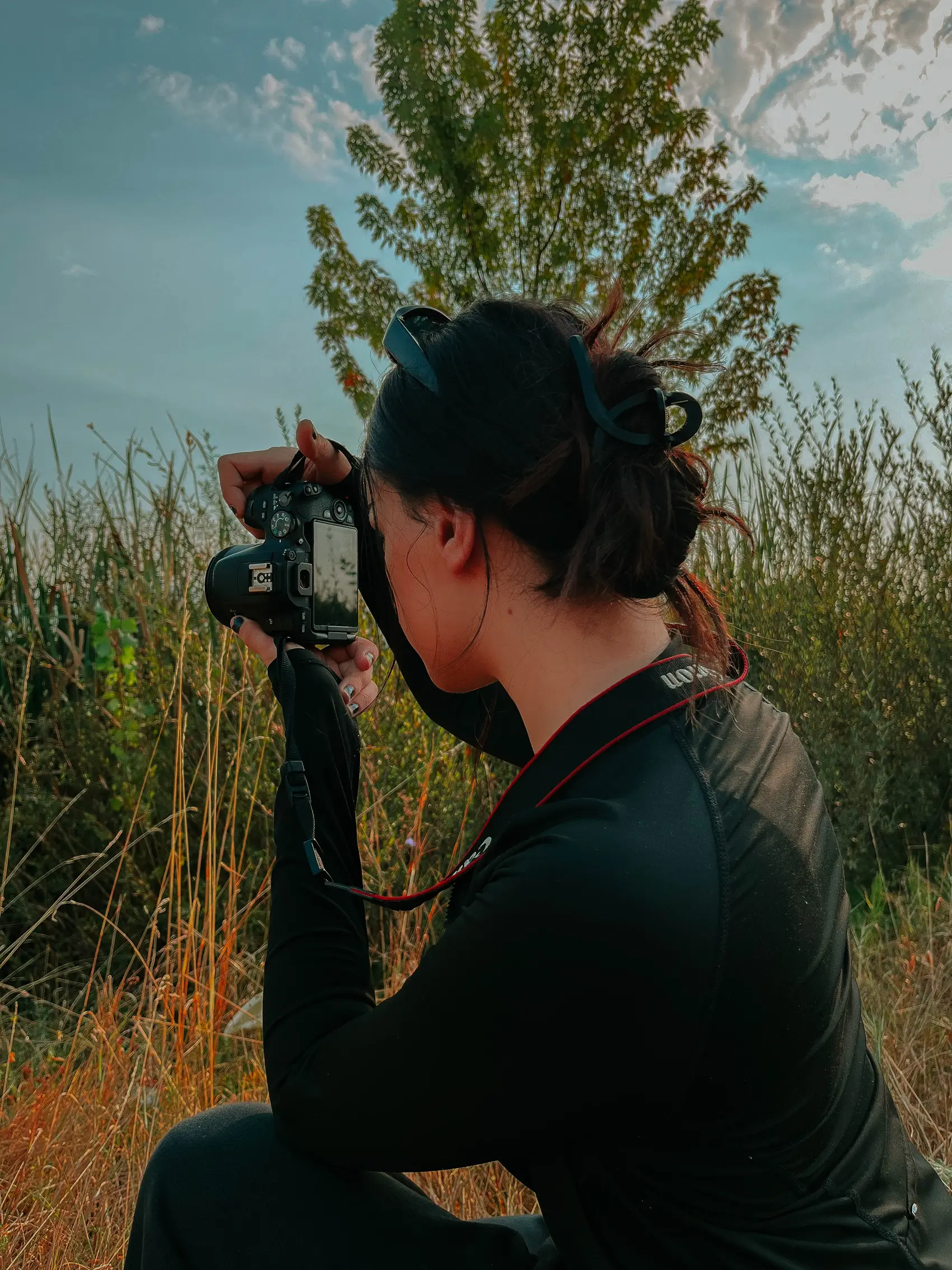 Engjellushe Avdiu kneeling in tall grass, framing a shot with her camera